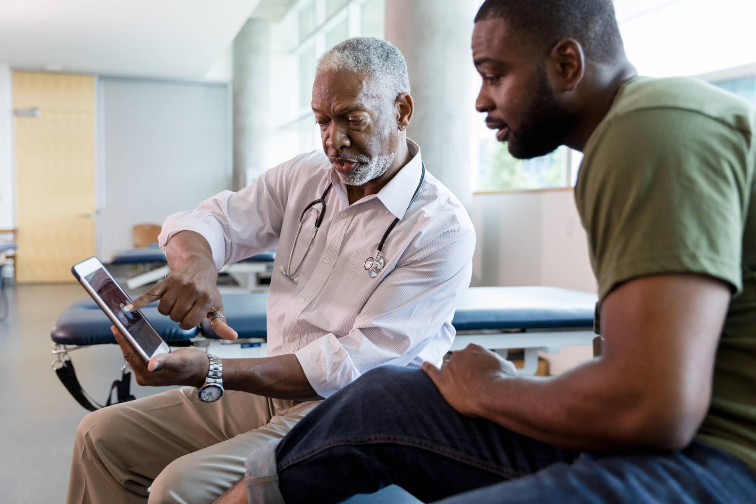 The mid adult male patient looks with interest as the mature adult male doctor points to the x-rays on the digital tablet screen.