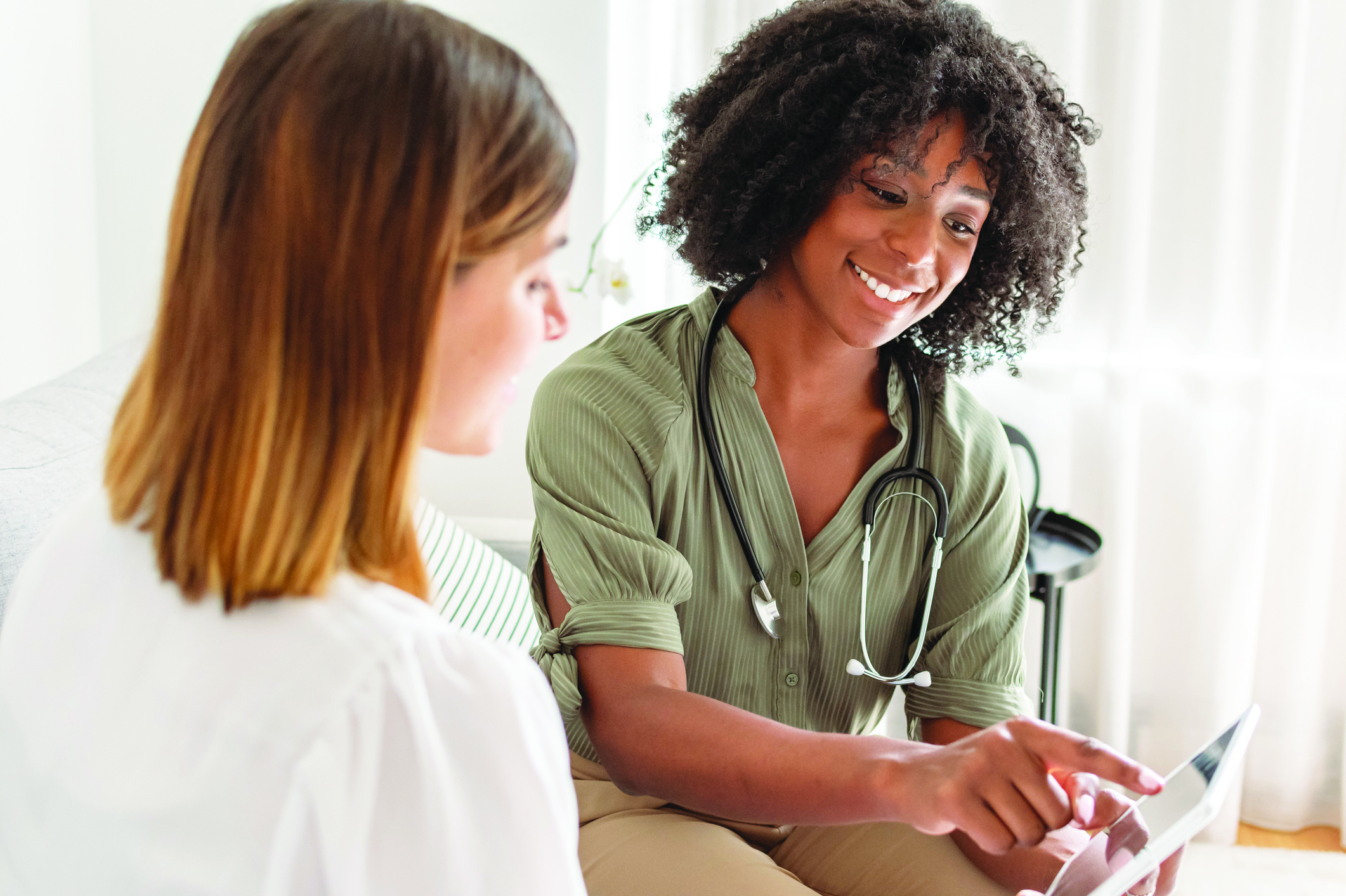 A young female doctor of African American descent smiles while reviewing test results on a tablet computer with a female patient during a medical appointment