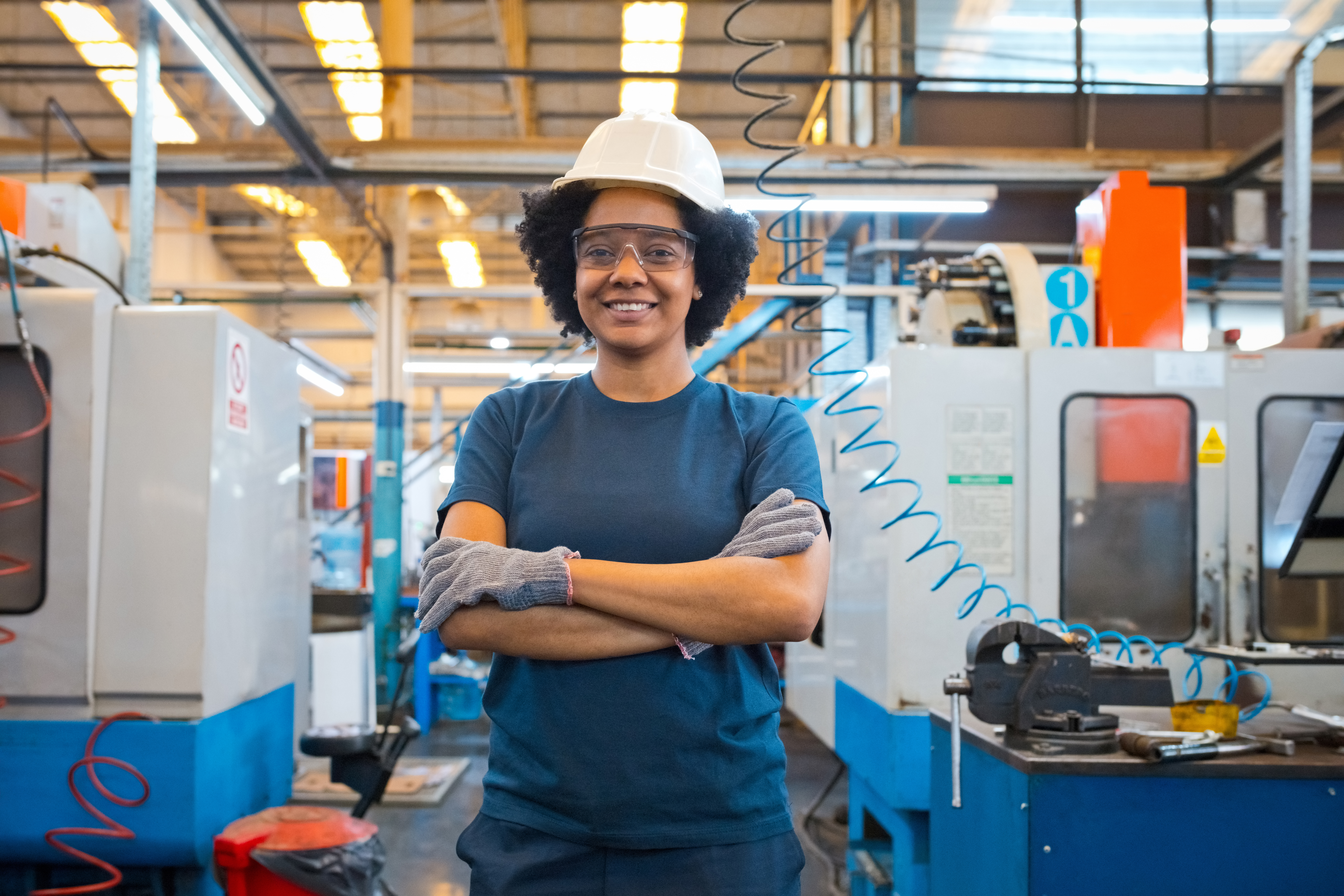Portrait of smiling female blue-collar worker wearing protective glasses and work helmet standing with arms crossed in industry.